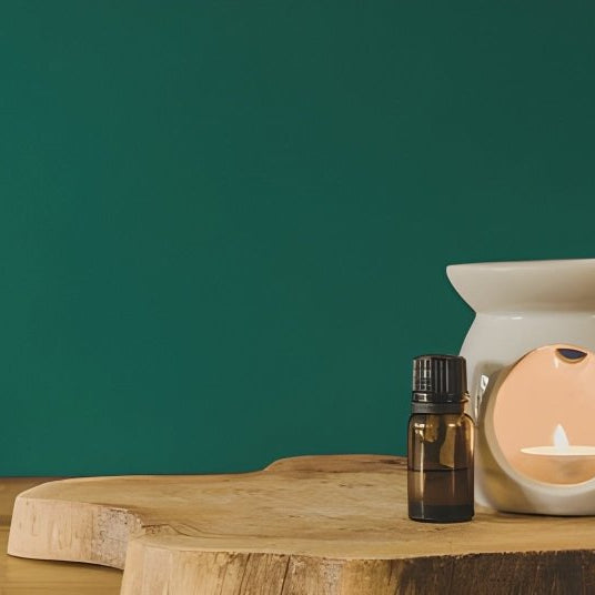 White ceramic tealight wax melt burner on a natural wooden slab with an amber fragrance bottle, styled against a deep green backdrop.