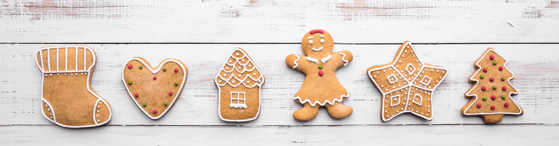 Row of iced gingerbread biscuits—stocking, heart, house, gingerbread person, star and Christmas tree—on a white wooden background, evoking spiced festive baking.