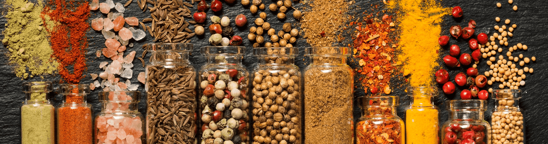 Overhead of glass jars filled with mixed peppercorns, cumin, coriander, mustard seeds and pink salt, with turmeric and chilli scattered on dark slate—evoking warm, spicy-woody fragrance.