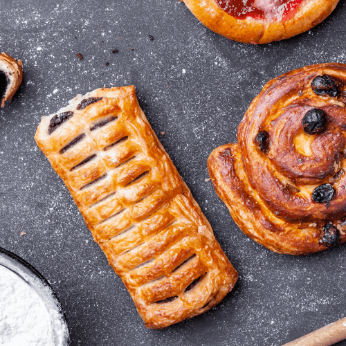 Overhead shot of assorted pastries—croissants, cinnamon roll, strudel and iced buns—on a dark slate surface dusted with flour, evoking vanilla-caramel sweetness.