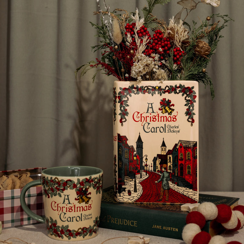 Large A Christmas Carol ceramic book vase filled with winter foliage; mug and holiday décor nearby