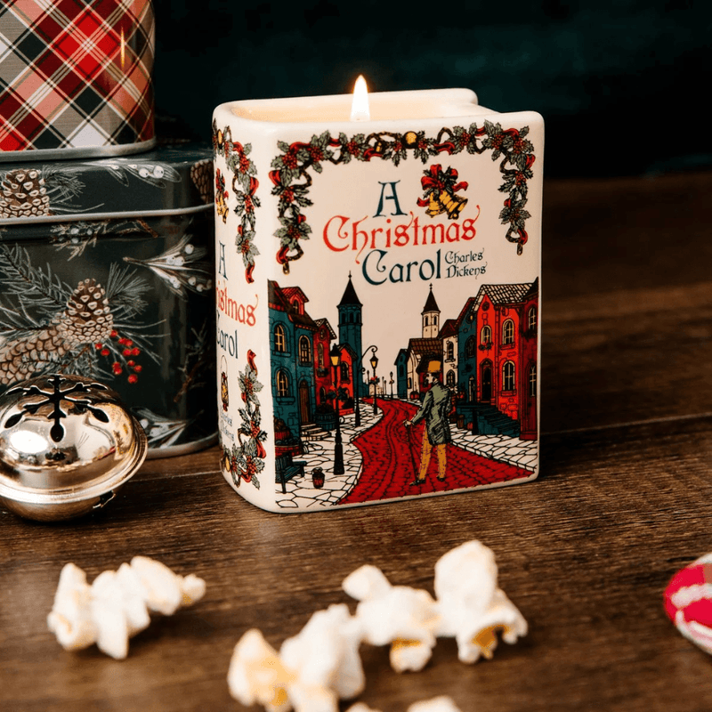 Lit A Christmas Carol ceramic book candle on a wooden table beside Christmas tins, a silver bell and festive decorations
