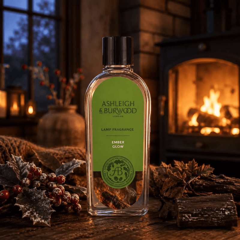 Ashleigh & Burwood Ember Glow Lamp Fragrance displayed by a glowing fireplace with frosted holly berries and logs