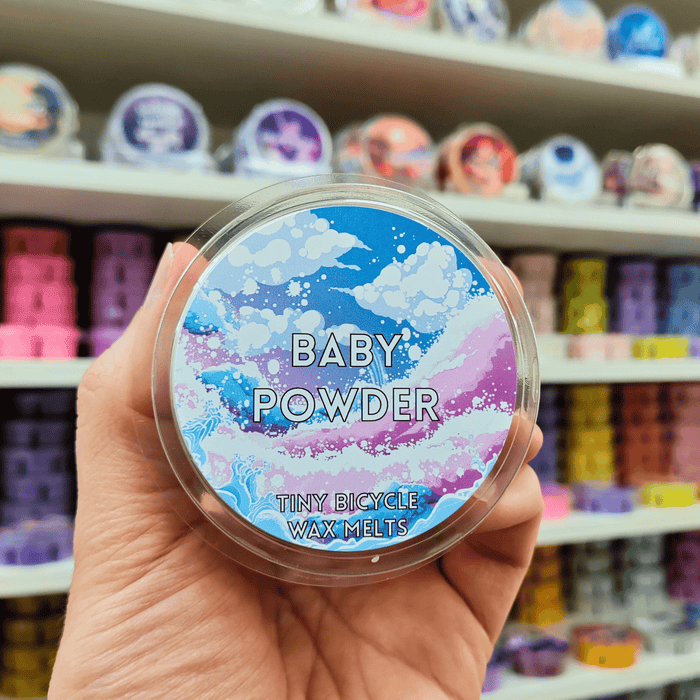 Baby Powder wax melt pot held in hand with pastel label, lifestyle image with wax melts on shelves in the background