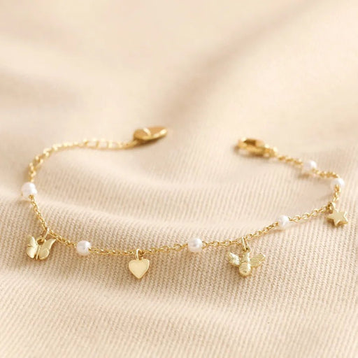 Close-up of a gold-tone chain bracelet with tiny pearls and charms—a butterfly, heart, bee and star—styled on soft cream fabric.