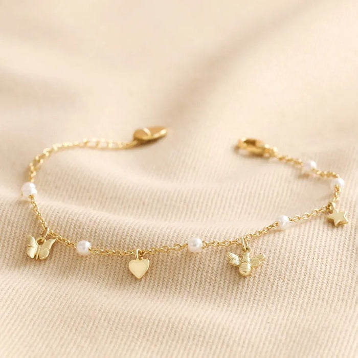 Close-up of a gold-tone chain bracelet with tiny pearls and charms—a butterfly, heart, bee and star—styled on soft cream fabric.