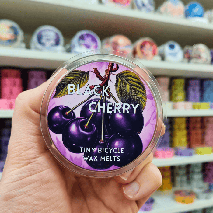 Black Cherry wax melt pot held in hand with illustrated cherry label, lifestyle image with colourful wax melts displayed on shelves in the background