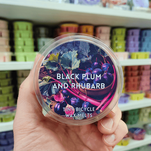 Black Plum and Rhubarb wax melt pot held in hand with illustrated fruit label, lifestyle image with colourful wax melts on shelves in the background