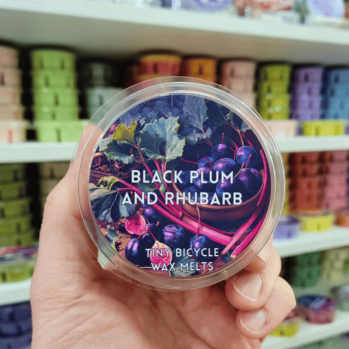 Black Plum and Rhubarb wax melt pot held in hand with illustrated fruit label, lifestyle image with colourful wax melts on shelves in the background