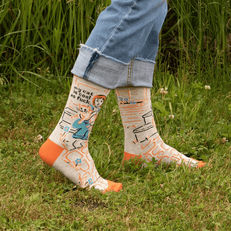 Blue Q women’s crew socks in orange and teal with retro cat graphic and text my cat is cool as fuck, model standing on grass
