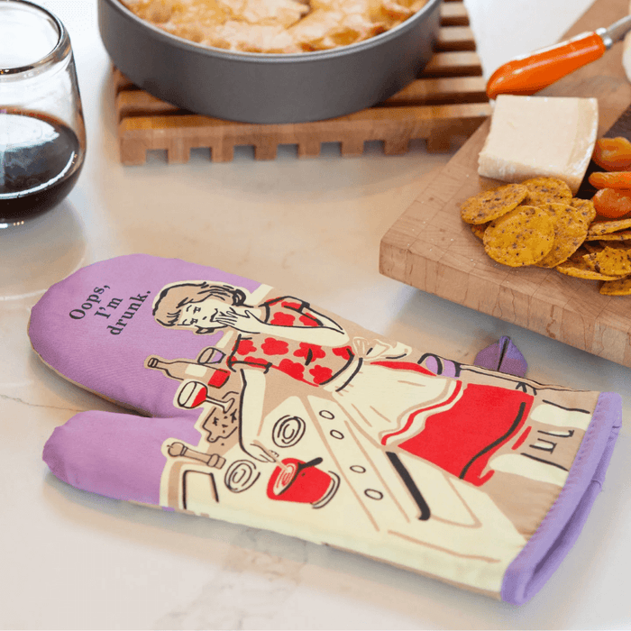 Blue Q Oops I’m Drunk oven mitt on marble beside cheese board, crackers and red wine