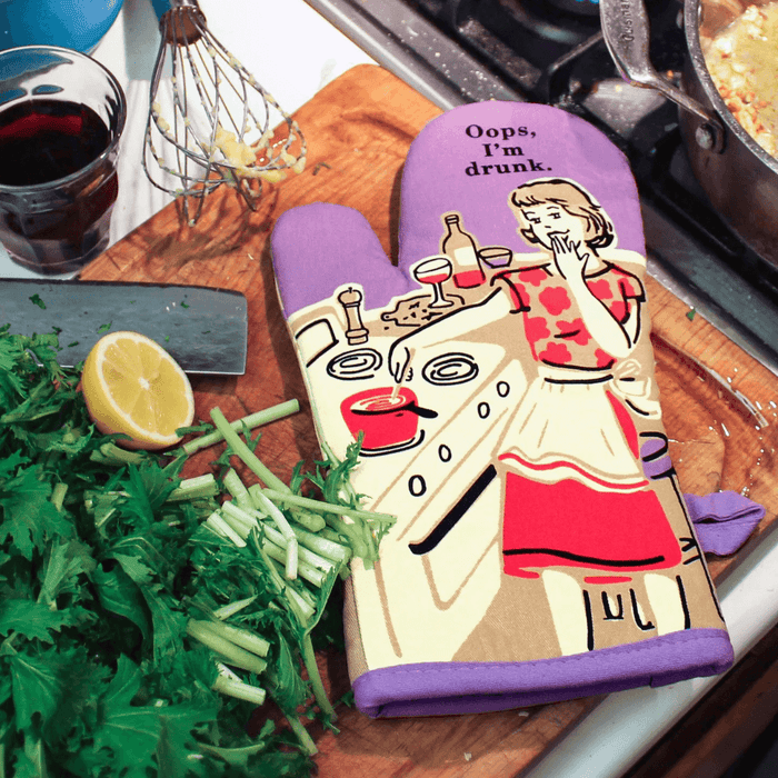 Blue Q Oops I’m Drunk oven mitt on chopping board with herbs and lemon by the hob


