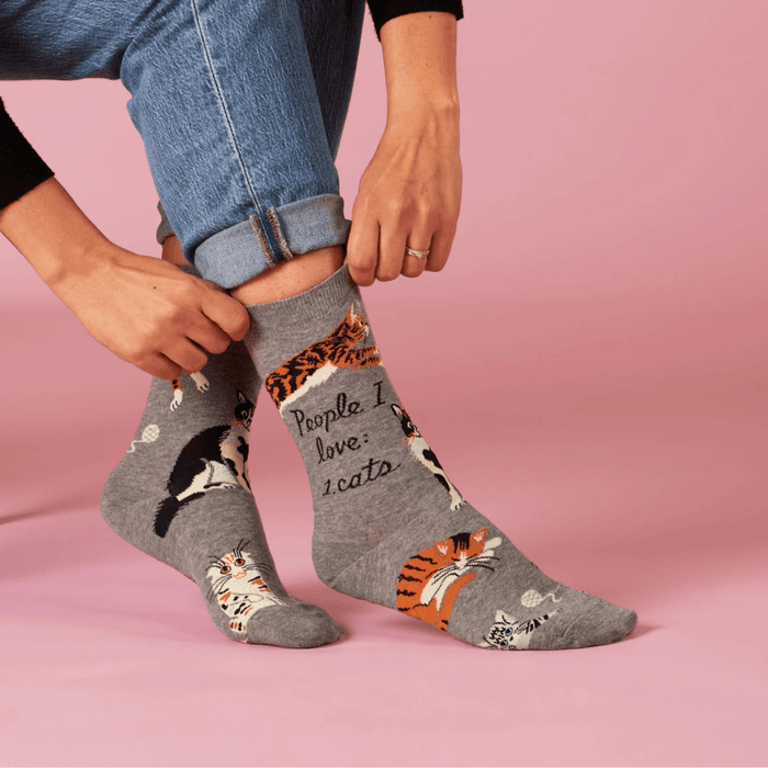 Hands adjusting Blue Q People I Love: Cats grey crew socks with illustrated cats, studio pink background