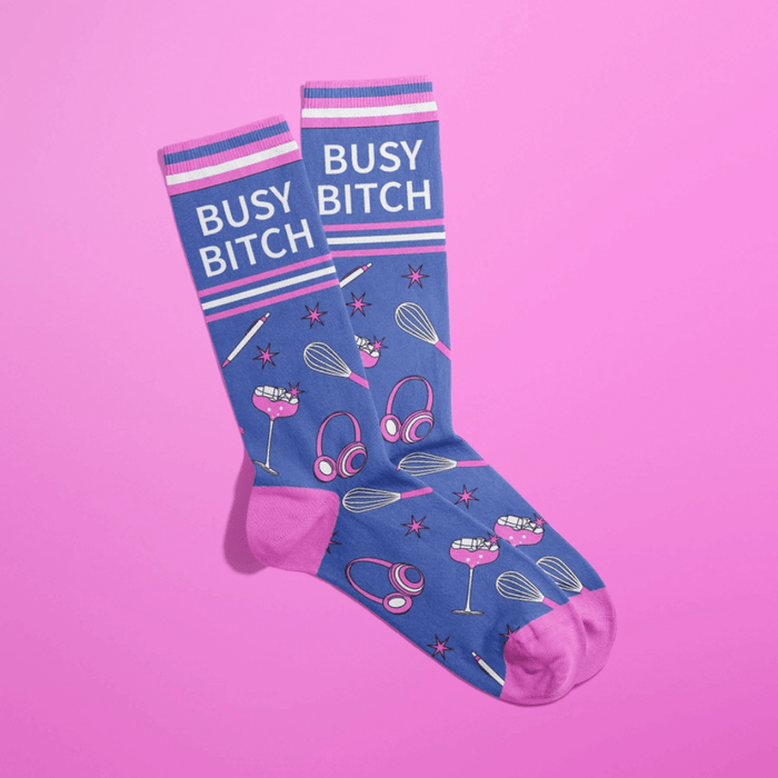 Overlapped blue women’s crew socks with pink heels and toes, Busy Bitch slogan, cocktails, headphones, whisks and stars on a pink backdrop.
