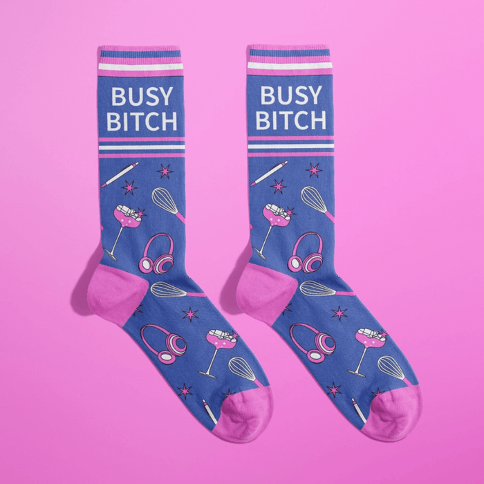 Pair of blue women’s novelty crew socks with pink accents and Busy Bitch text, sprinkled with cocktail glasses, headphones, whisks and stars.