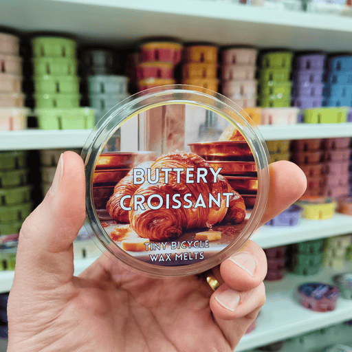 Buttery Croissant wax melt pot held in hand with croissant label, lifestyle image with colourful wax melts on shelves in the background
