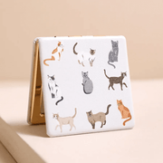 Closed square compact mirror with grey and ginger cat print on a white PU cover and gold-tone edges.