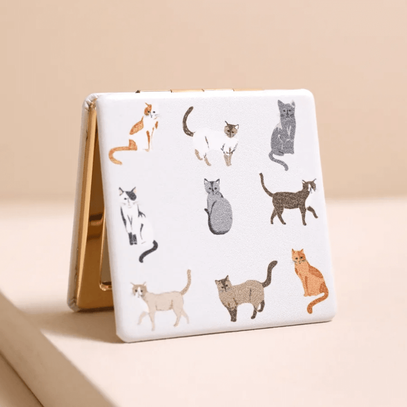 Closed square compact mirror with grey and ginger cat print on a white PU cover and gold-tone edges.