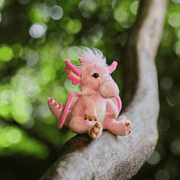 Charlie Bears Festoon dragon plush soft toy perched on a tree branch outdoors, pink wings and fluffy white mane