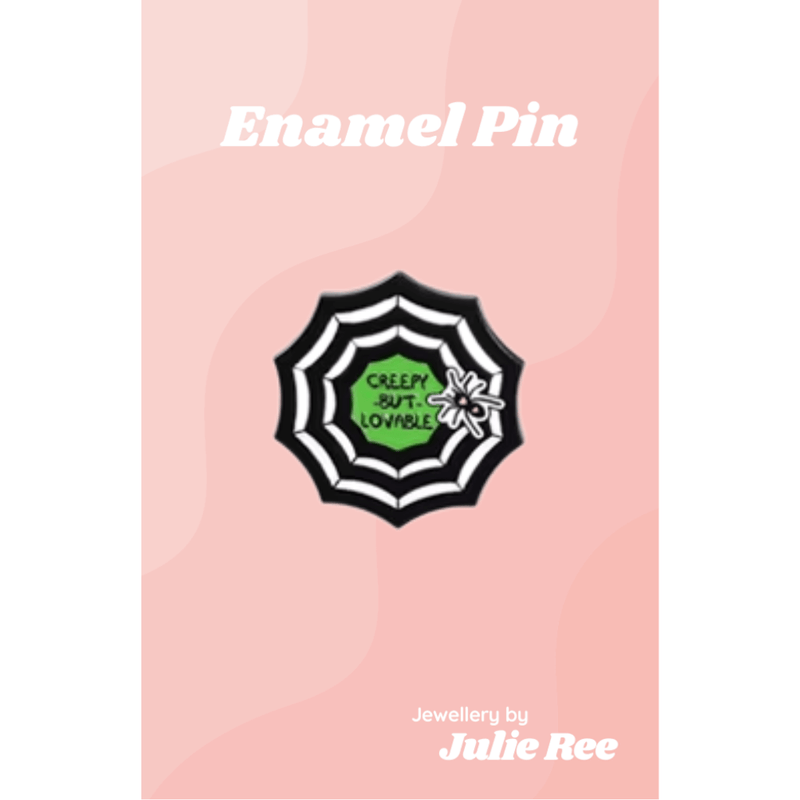 Black and white spiderweb enamel pin with a small spider and green centre reading Creepy But Lovable, on a pink backing card.