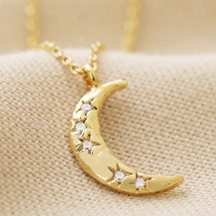 Close-up of a gold crescent moon pendant dotted with tiny crystal stars on a dainty chain, celestial necklace on neutral fabric.