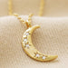 Close-up of a gold crescent moon pendant dotted with tiny crystal stars on a dainty chain, celestial necklace on neutral fabric.