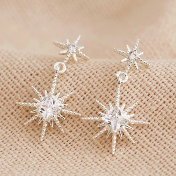Close-up of silver double star drop earrings with faceted crystals on neutral fabric.
