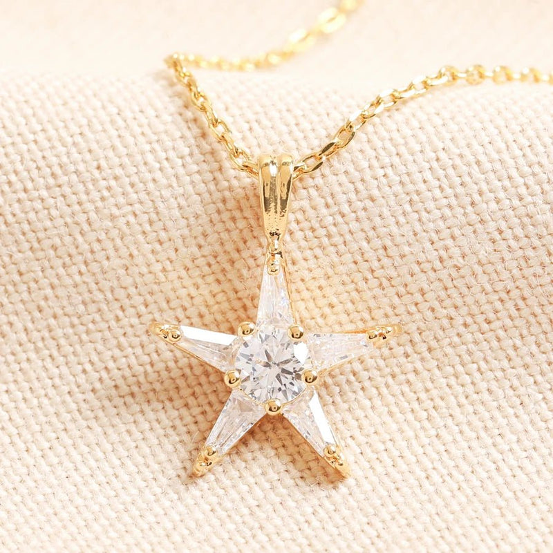 Gold star pendant necklace set with clear crystals on a fine chain, close-up of celestial jewellery piece on neutral fabric.