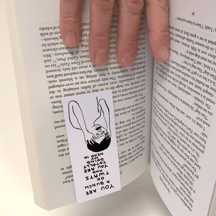 Bookmark gripping pages as a hand turns the book; Shrigley artwork visible upside-down between sheets.


