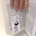 Bookmark gripping pages as a hand turns the book; Shrigley artwork visible upside-down between sheets.

