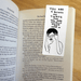 Magnetic bookmark clipped inside an open novel, showing Shrigley’s stressed figure and rude slogan.
