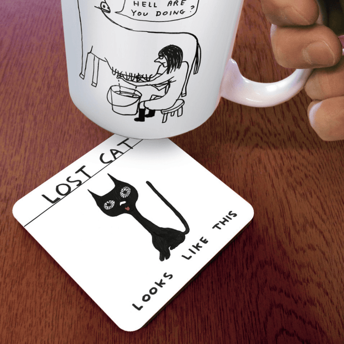 Lost Cat coaster on wooden table under illustrated mug; quirky black cat design visible at an angle.
