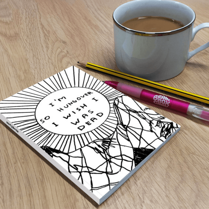 Lifestyle image: humorous hungover notebook on wooden desk beside mug of tea, pencil and pink pen.