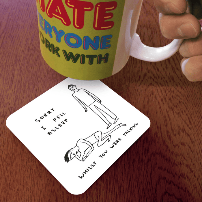Sorry I Fell Asleep coaster on wooden table beneath illustrated mug; sleepy design visible at an angle.

