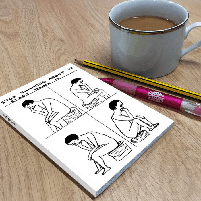 Lifestyle image: humorous motivational notebook on wooden desk beside mug of tea, pencil and pink pen.

