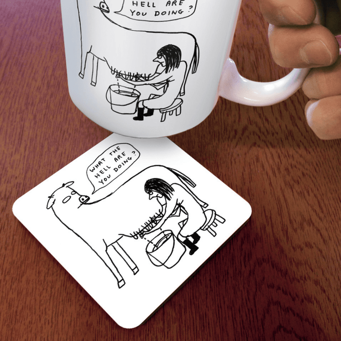 What the Hell coaster on wooden desk beneath illustrated mug; humorous cow-and-milker design visible at an angle.

