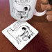 What the Hell coaster on wooden desk beneath illustrated mug; humorous cow-and-milker design visible at an angle.

