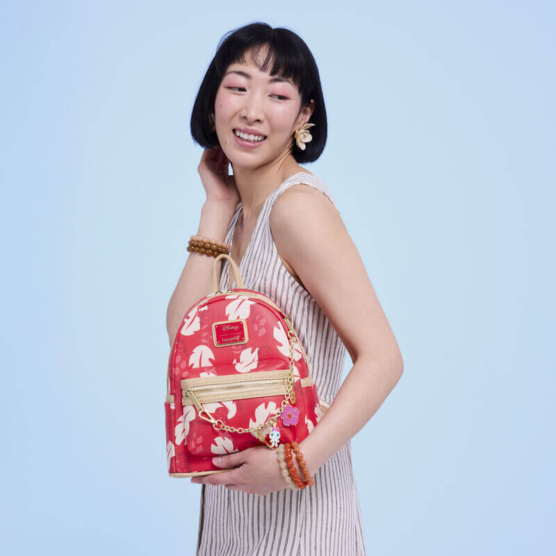 Model posing with red Loungefly Disney Lilo & Stitch Back Pack bag; cream monstera leaves, gold zips and tropical charm chain.	