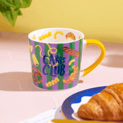 Colourful Eleanor Bowmer Carbs Club coffee mug beside a croissant