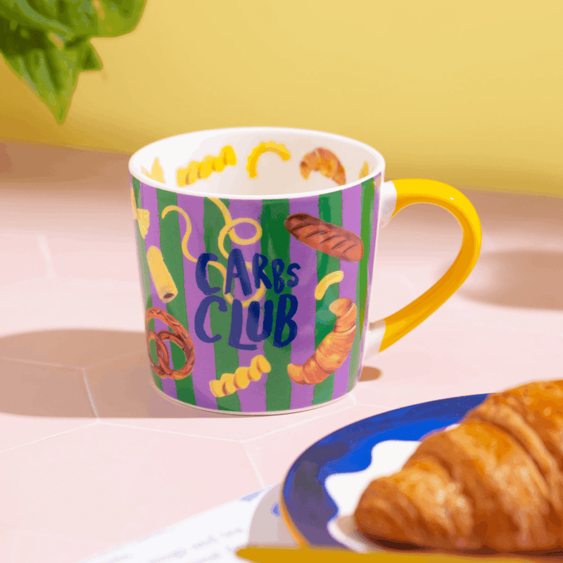 Colourful Eleanor Bowmer Carbs Club coffee mug beside a croissant