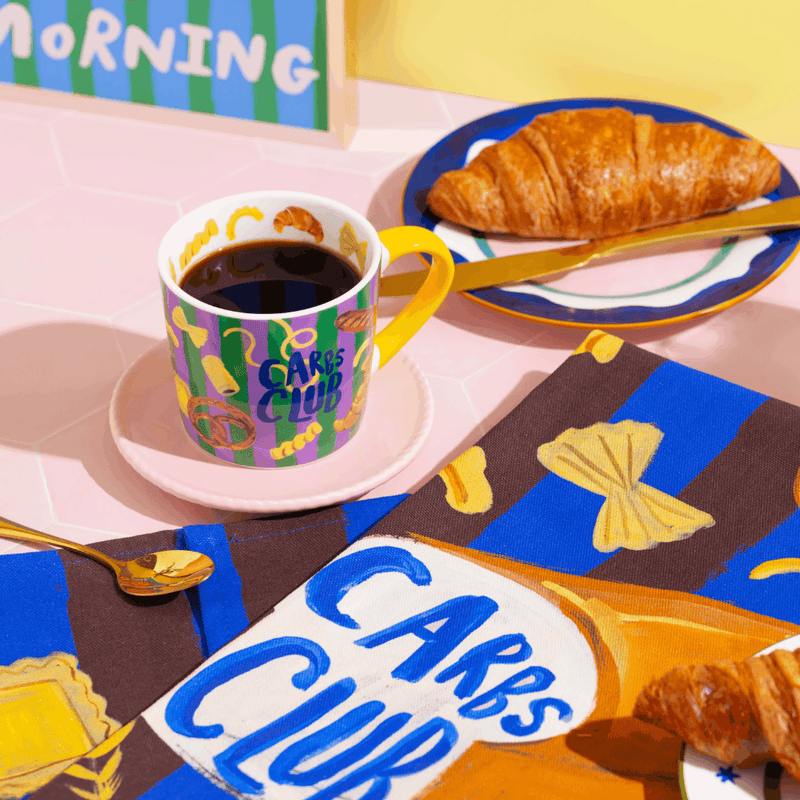 Eleanor Bowmer Carbs Club mug styled with croissants and colourful breakfast accessories
