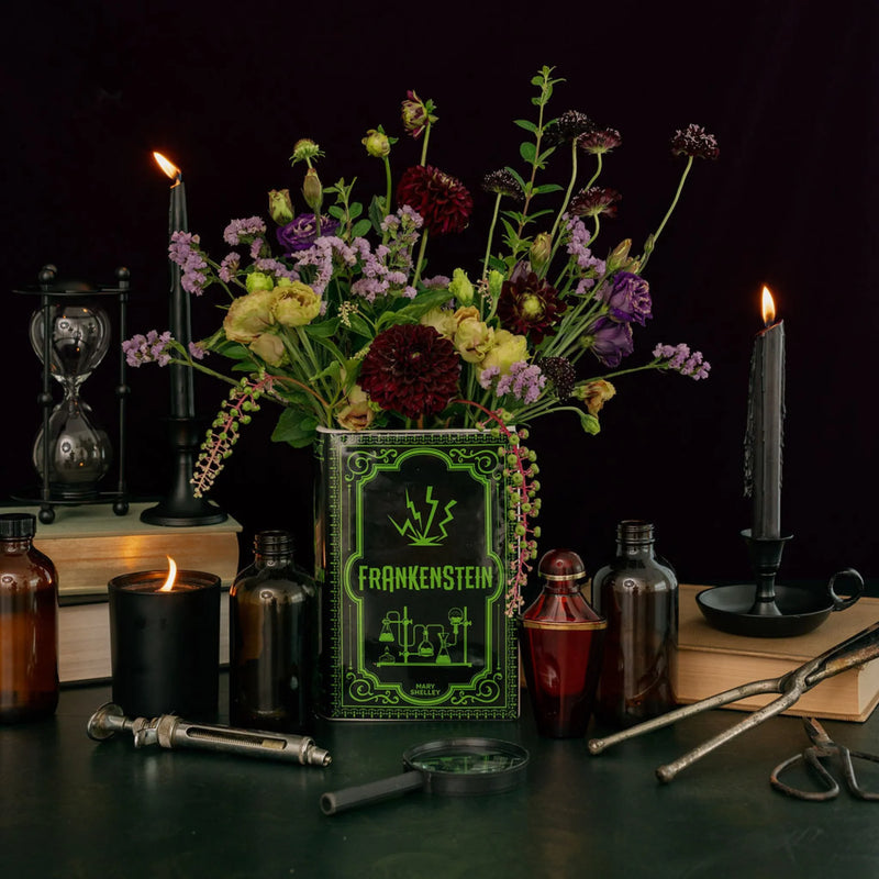 Frankenstein book vase filled with flowers on a gothic tabletop with candles and apothecary bottles.