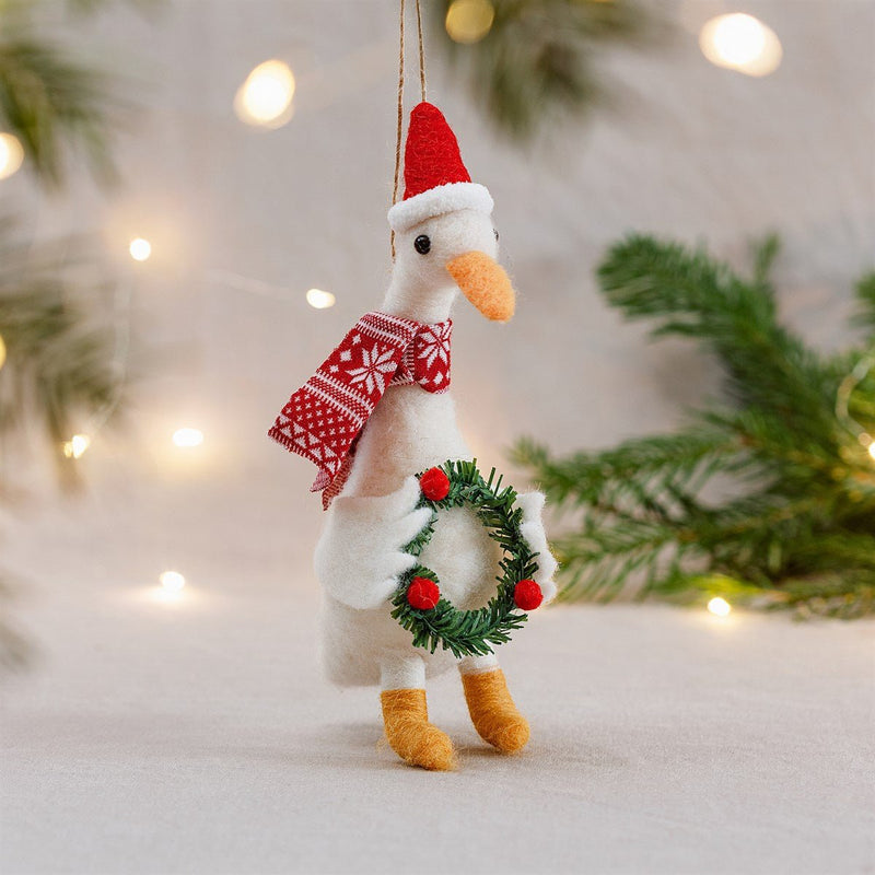 Fabric goose ornament with Santa hat and wreath hanging on twine among fairy lights and fir branches in a cosy Christmas scene.