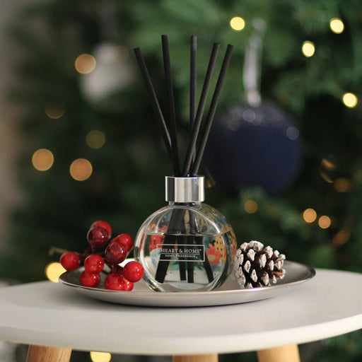 Round glass reed diffuser with black reeds on a tray by a Christmas tree, styled with berries and a pine cone, soft festive lights in background.