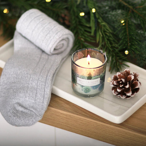Lit Heart & Home Christmas Tree mini candle beside rolled grey socks on a tray with pinecone and festive greenery, soft fairy lights in background.