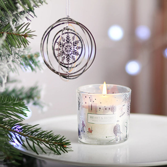 Silver spinning tree decoration hanging beside a lit Heart & Home Snowflake Sparkle mini candle in a frosty village glass, soft lights in background.
