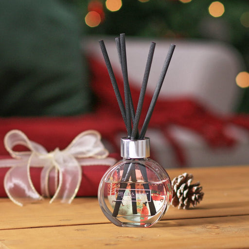 Round bauble-style reed diffuser with chunky black reeds on a wooden table, Christmas tree bokeh in the background and ribboned gift beside it.