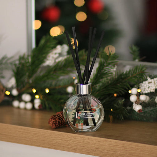Round bauble-style reed diffuser with chunky black reeds on a mantle styled with winter greenery and lights; soft festive bokeh in the background.