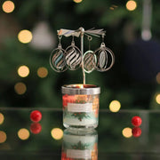 Lit Heart & Home mini candle with silver carousel topper; bauble-shaped charms spin above the flame on a festive, bokeh-lit background.