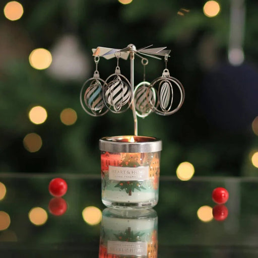 Lit Heart & Home mini candle with silver carousel topper; bauble-shaped charms spin above the flame on a festive, bokeh-lit background.